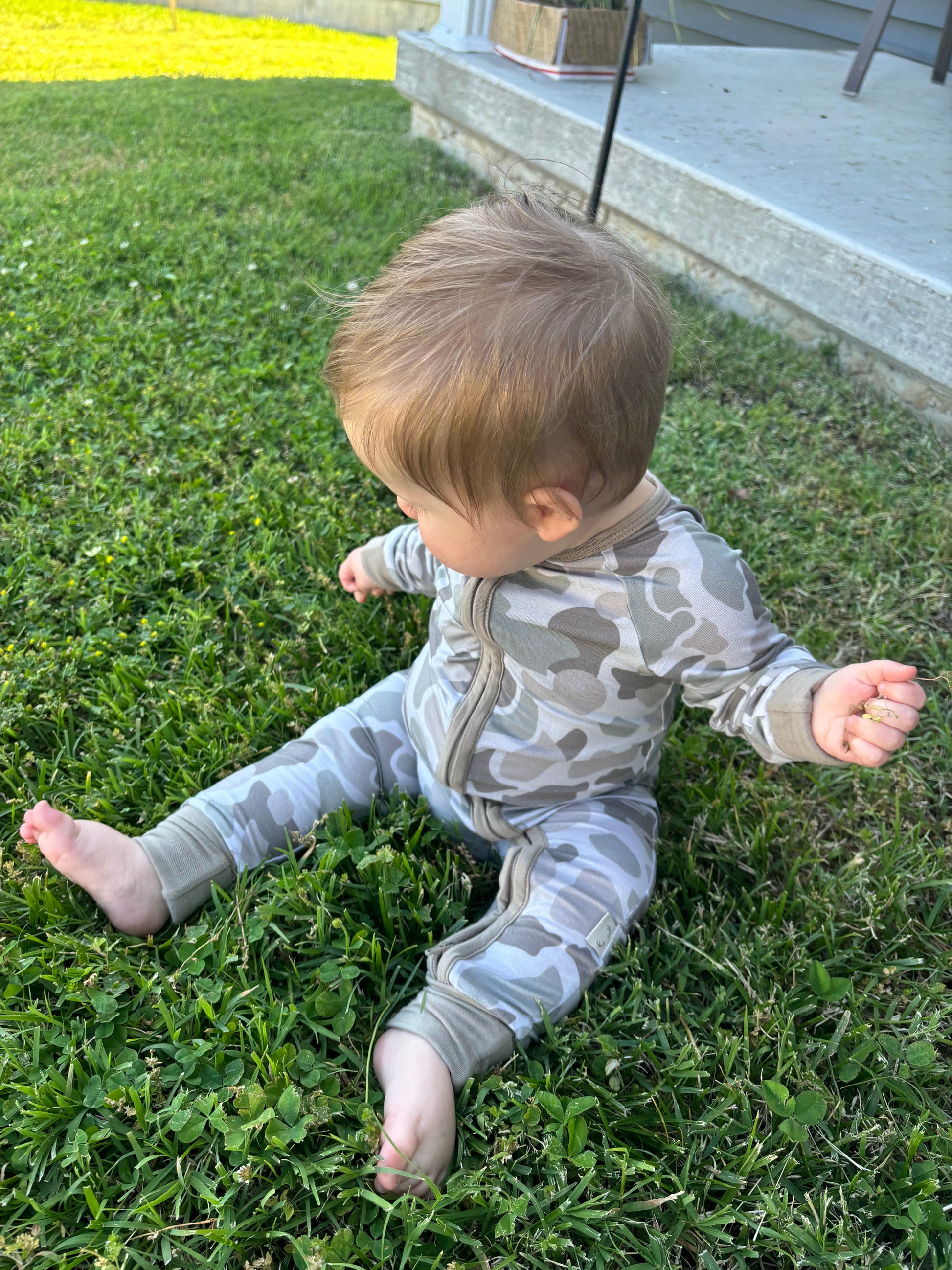 Baby wearing Button Buck Camo Baby Zippy, a soft bamboo one-piece outfit, sitting on grass.